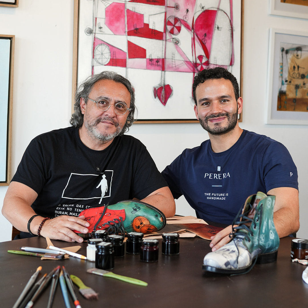 Two men sitting at a table with art supplies and colorful shoes, surrounded by framed artwork.